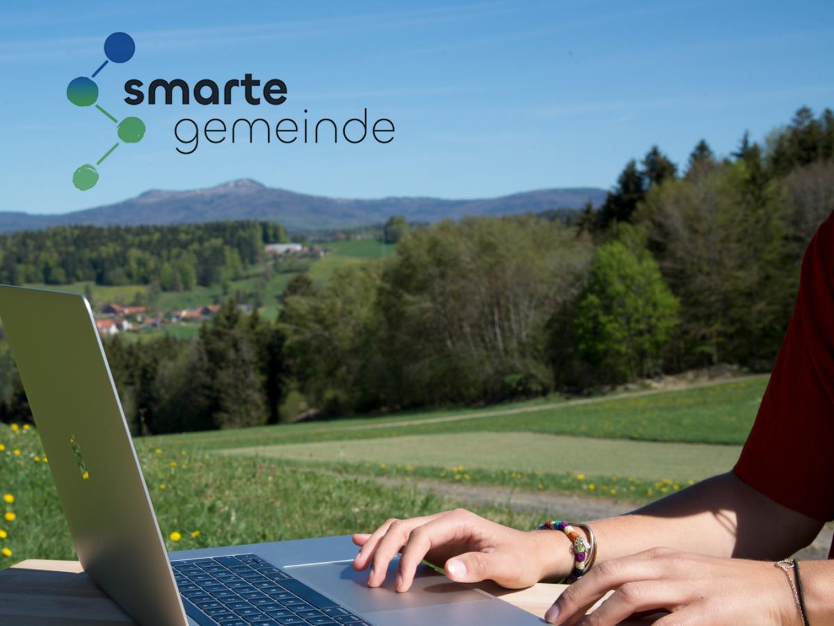 Person arbeitet an Laptop auf Holztisch in bayerischer Alpenlandschaft mit Bergen, Wiesen und Bauernhäusern.