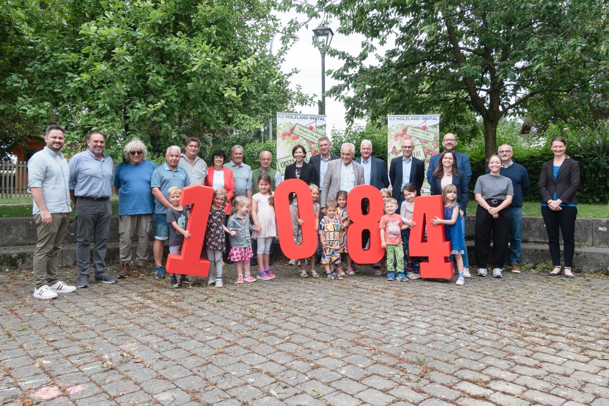 Eine Gruppe von Personen (Erwachsene und Kinder) halten die große rote Zahl 1084 hoch.