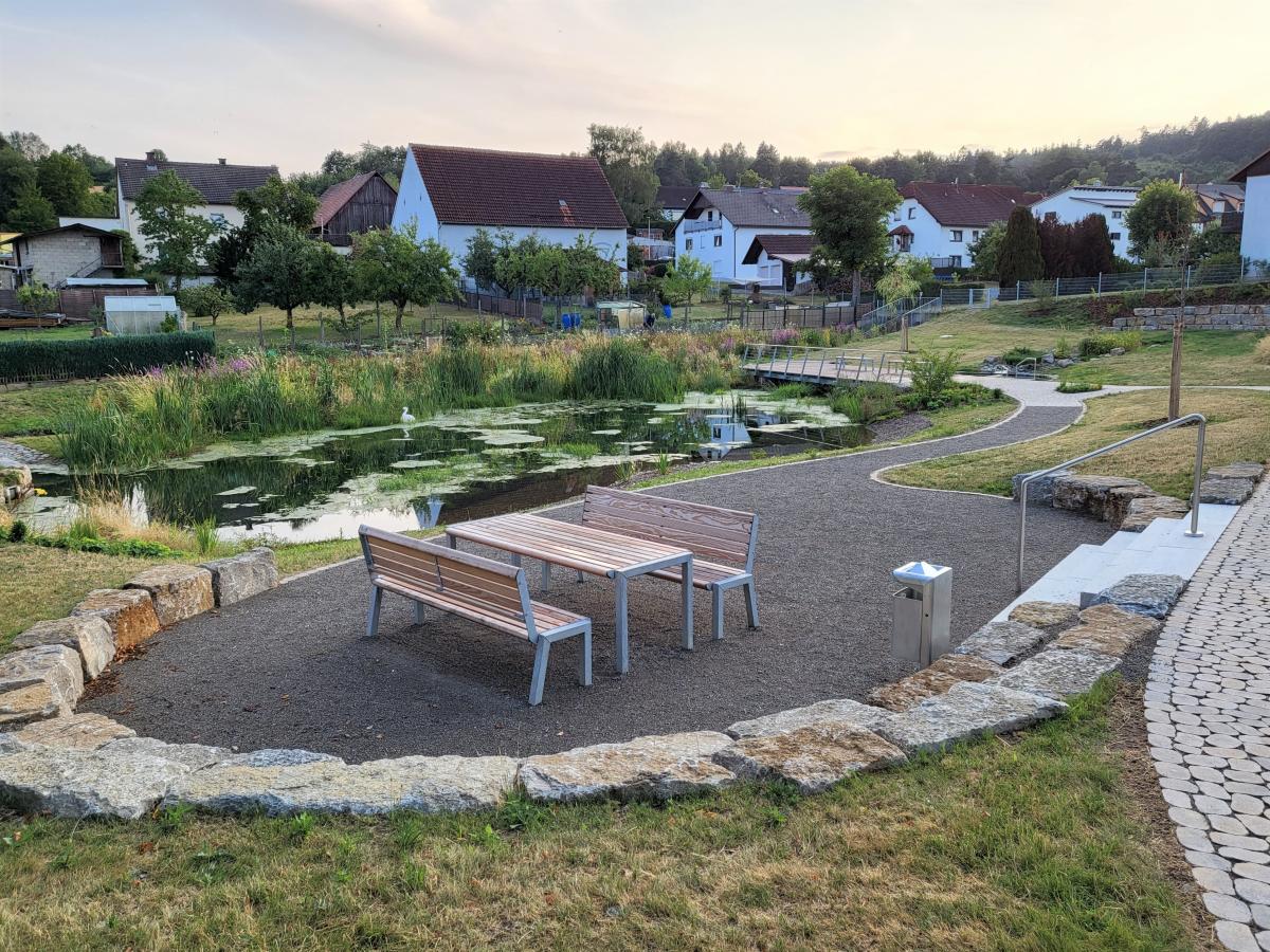 Vor dem Ortssaum von Waldfenster schmiegt sich ein kleiner See in den sanften Abhang. Davor steht eine Holzsitzgruppe auf einer dunkel geschotterten Fläche für den gemütlichen Aufenthalt. Am rechten Bildrand ist ein Wassertretbecken zu erkennen.