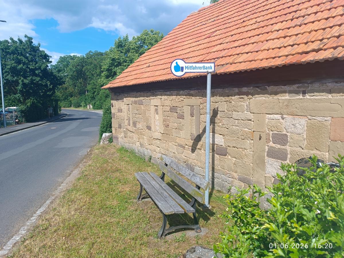 Am Straßenrand ist eine Haltestelle mit Fahrbank vor einem Stadel eingrichtet worden.