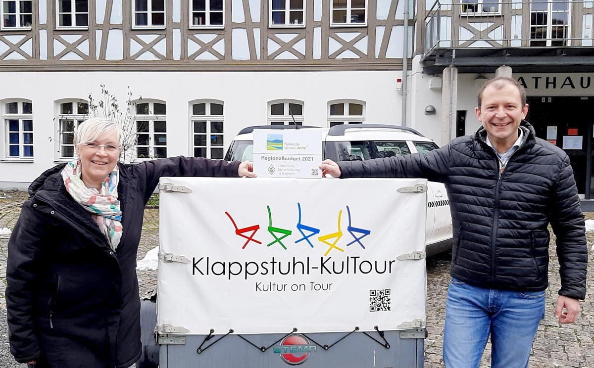 Bürgermeister Marco Trautner übergibt Förderschild an eine Frau vor dem Anhänger der Klappstuhl-KulTour