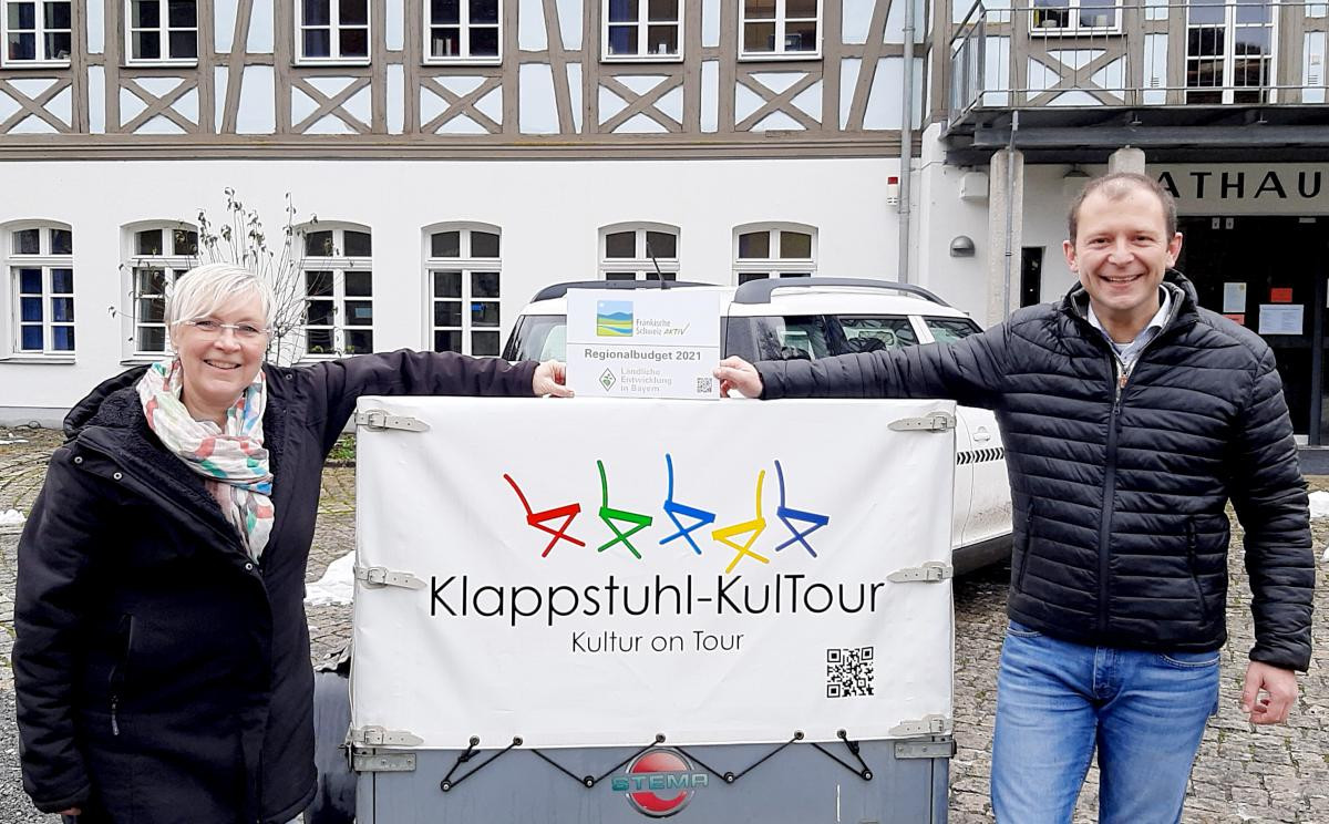 Bürgermeister Marco Trautner übergibt Förderschild an eine Frau vor dem Anhänger der Klappstuhl-KulTour