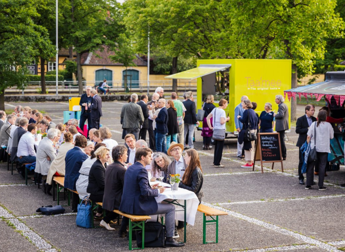 Auf einem Parkplatz sitzen mehrere Personen einer langen Tafel aus Biertischgarnituren.
