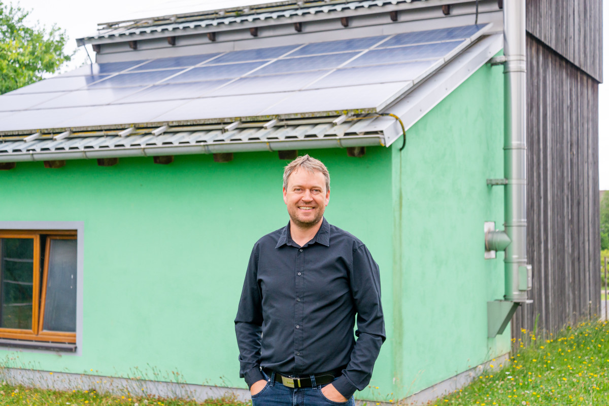 Das Bild zeigt Andreas Bodensteiner vor einem Gebäudeteil in dem Energie für die Gemeinde produziert wird. Das türkischgrün gestrichene Gebäude hat eine Solaranlage auf dem Dach.