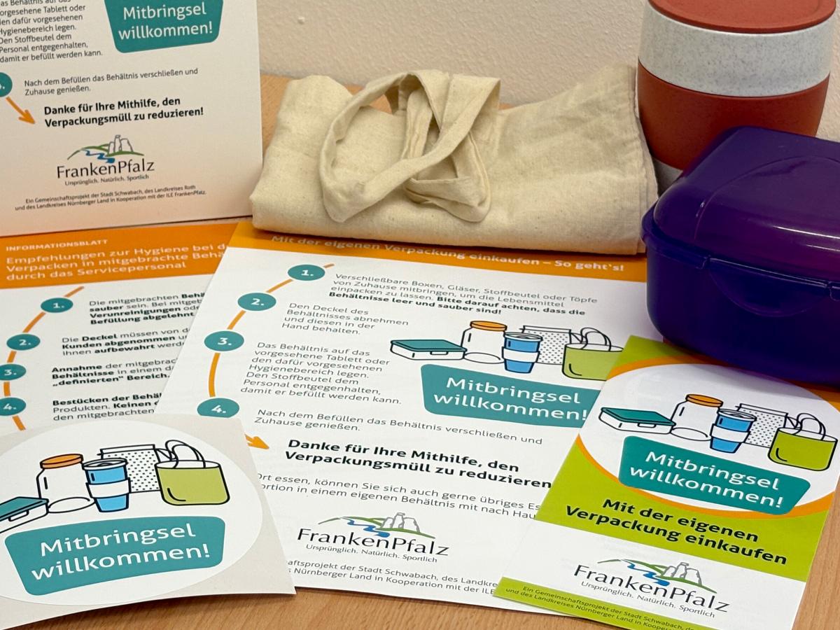 Aufkleber und Infoblätter informieren über die Aktion - sie liegen auf einem Tisch. Brotboxen, ToGo Becher und Jutetaschen flankieren die Printmaterialien