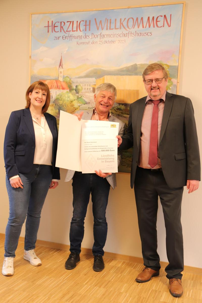Zwei Herren und eine Frau stehen vor einem Gemälde der Ortsmitte von Kumreut. Der Mann in der Mitte hält einen Förderbescheid in seinen Händen.