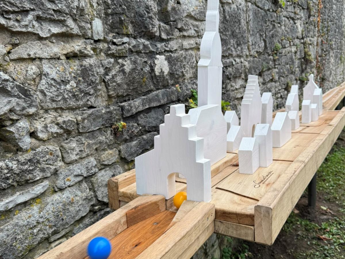 Eine blaue und eine gelbe Kugel liefern sich in einer hölzernen Bahn ein Wettrennen. Die Strecke zieht sich an der Dorfmauer entlang und ist überbaut mit stilisierten Gebäuden.