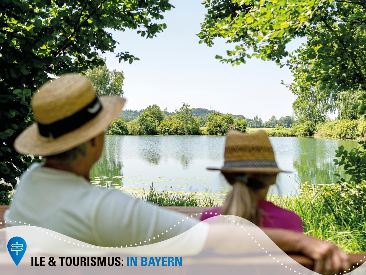 Eine ältere Dame  und ein Mädchen mit Strohhüten sitzen auf einer Bank und blicken auf einen See.